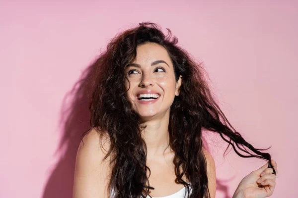 Positive brunette woman touching curly hair on pink background — Stock Photo