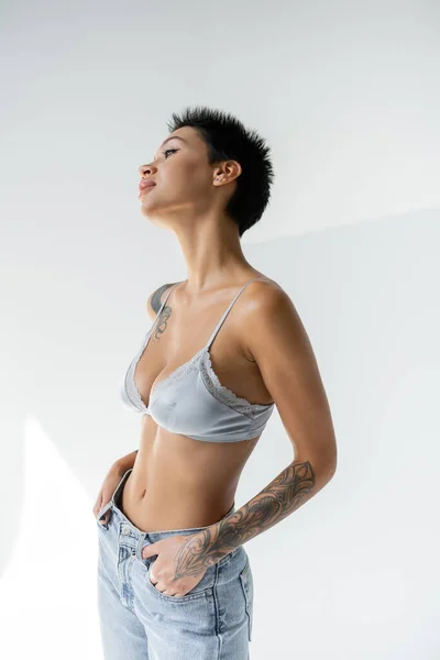 Seductive tattooed woman in silk bra posing with hands in pockets of jeans on grey background — Fotografia de Stock