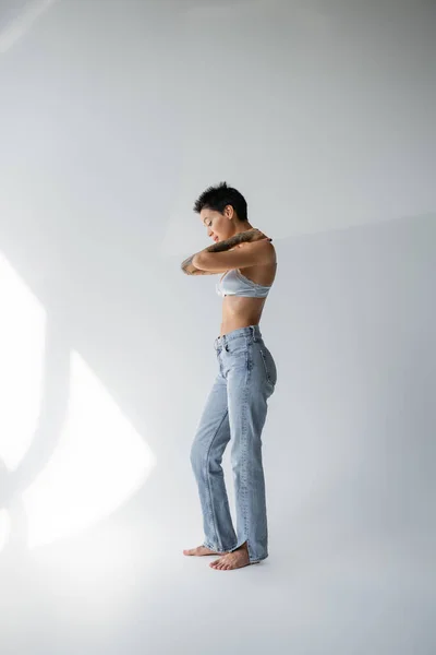 Full length of barefoot tattooed woman in blue jeans and bra standing with hands on shoulders on grey background — Fotografia de Stock