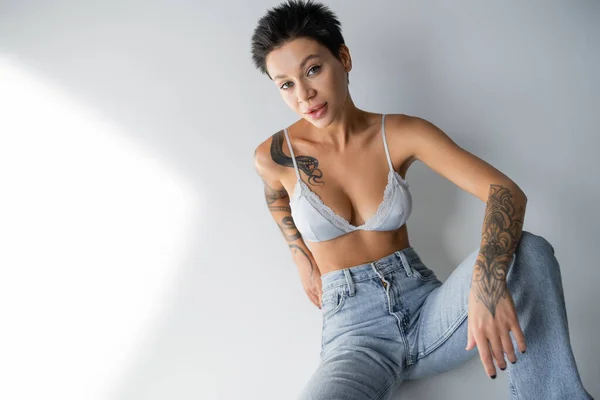 Brunette woman with sexy tattooed body sitting in jeans and bra on grey background — Fotografia de Stock