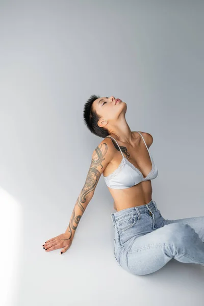 Seductive tattooed woman in silk bra and jeans sitting with closed eyes on grey background — Fotografia de Stock
