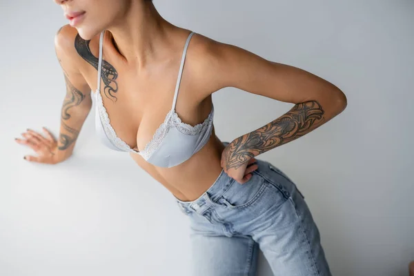 Vista parcial de mujer tatuada en jeans y sujetador posando con la mano sobre la cadera sobre fondo gris - foto de stock