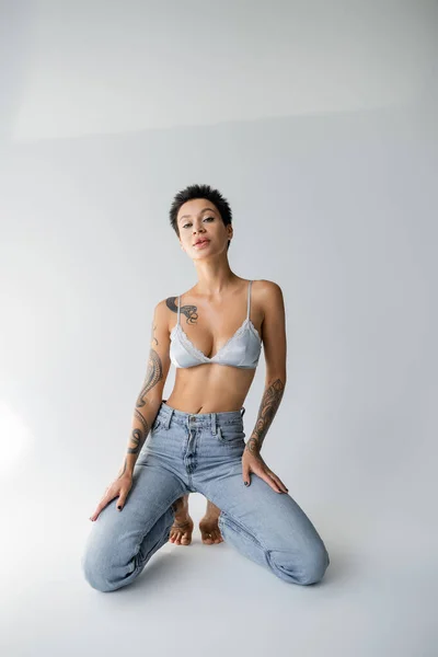 Sensual brunette woman in satin bralette and jeans posing on knees on grey background — Stockfoto