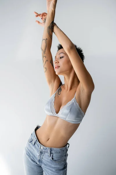 Tattooed and slender woman in silk bra and jeans standing with raised hands on grey background — Foto stock