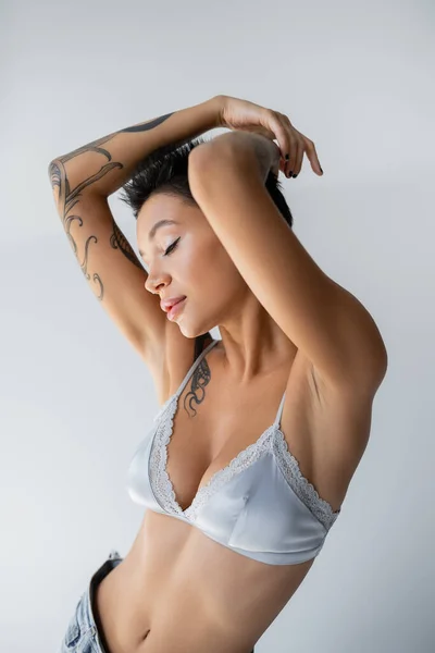 Sensual mujer tatuada en sujetador de seda posando con los ojos cerrados y las manos por encima de la cabeza aisladas en gris - foto de stock