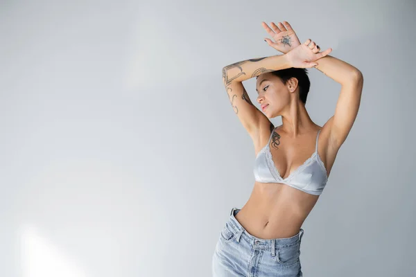 Young and slender woman in satin bra and jeans posing with tattooed hands above head on grey background — Fotografia de Stock
