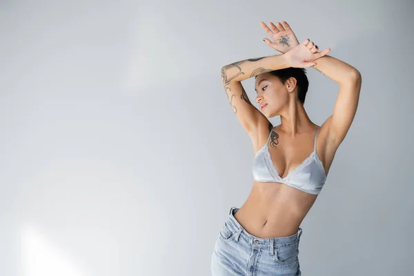 Mujer joven y delgada en sujetador de satén y jeans posando con las manos tatuadas por encima de la cabeza sobre fondo gris - foto de stock