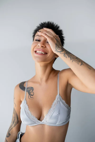 Cheerful tattooed woman in blue silk bra obscuring face with hand isolated on grey — Stock Photo