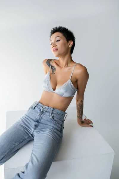 Young tattooed woman in blue jeans and satin bra posing on cube on grey background — Fotografia de Stock