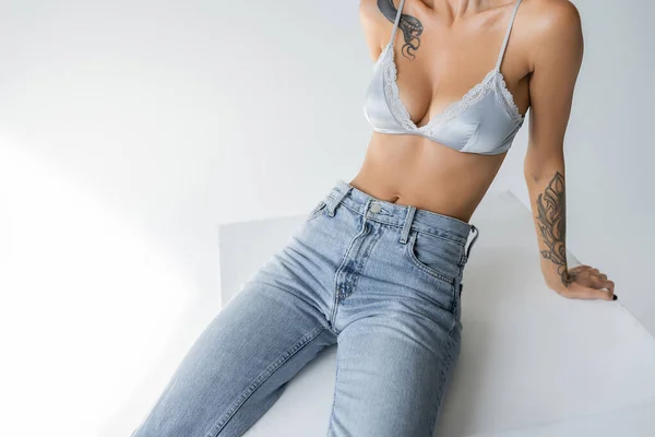 Partial view of slender woman in blue jeans and silk bralette sitting on cube on grey background - foto de stock