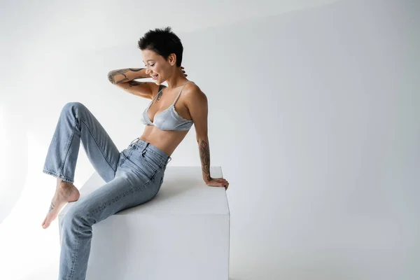 Smiling brunette woman in silk bra and jeans sitting on cube on grey background — Photo de stock