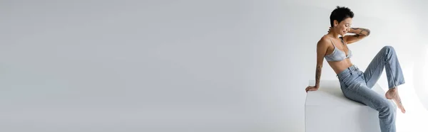 Smiling barefoot woman in bra and jeans sitting on cube on grey background, banner — Stock Photo