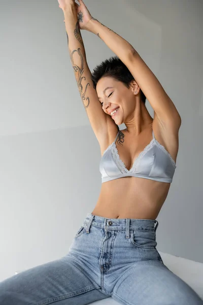 Joyful tattooed woman in jeans and silk bra sitting on cube with raised hands and closed eyes on grey background — Fotografia de Stock