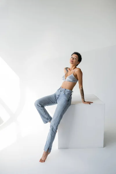 Full length of slender tattooed woman in blue jeans and silk bra looking at camera near cube on grey background — Photo de stock