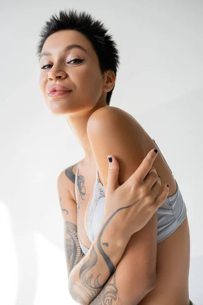 Sonriente mujer tatuada en sujetador azul posando con los brazos cruzados y mirando a la cámara sobre fondo gris - foto de stock