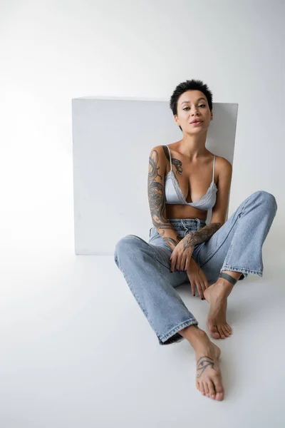Barefoot tattooed woman in blue jeans and bra sitting near cube and looking at camera on grey background — Fotografia de Stock