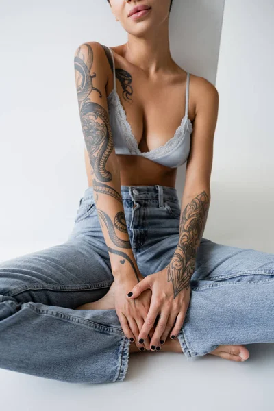 Visão parcial de mulher descalça tatuada em jeans e sutiã sentado com pernas cruzadas em fundo cinza — Fotografia de Stock