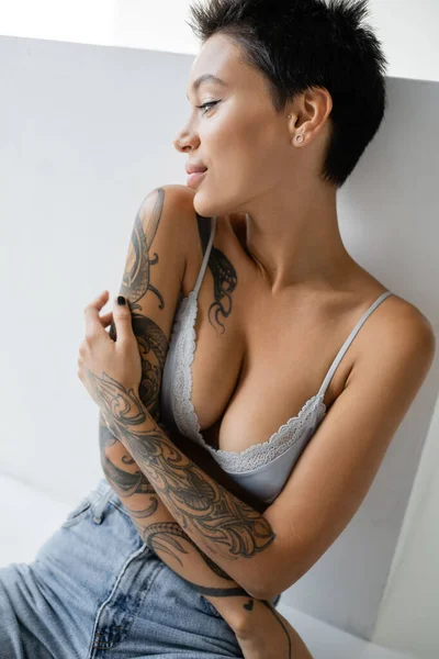 Positive tattooed woman with sexy body sitting in bra and jeans near cube on grey background — Photo de stock