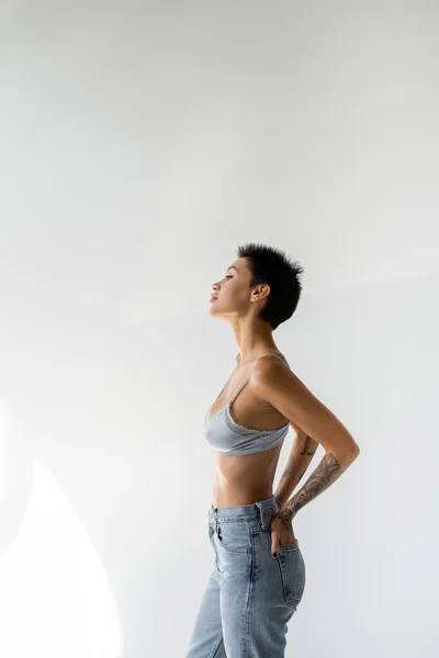 Side view of brunette short haired woman in blue bra standing with hands in back pockets of jeans on grey background — Photo de stock