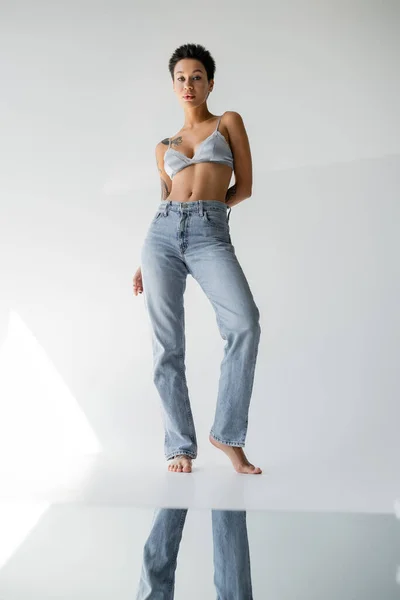 Full length of barefoot brunette woman in jeans and bralette standing near mirror on floor on grey background — Photo de stock