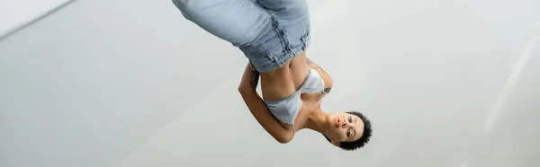 Mirror reflection of sexy woman in bralette and jeans standing with hands behind back on grey background, banner — Stock Photo