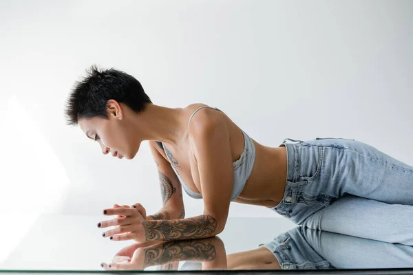 Side view of brunette short haired woman in blue jeans and bra lying on mirror on grey background — Stockfoto