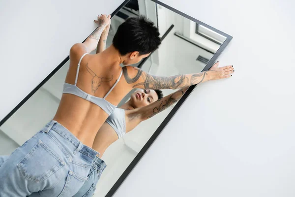 High angle view of seductive tattooed woman in bra and jeans lying on mirror on grey background — Stock Photo