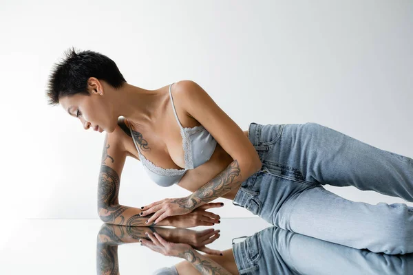 Mulher tatuada em bralete azul e jeans deitado no espelho em fundo cinza — Fotografia de Stock