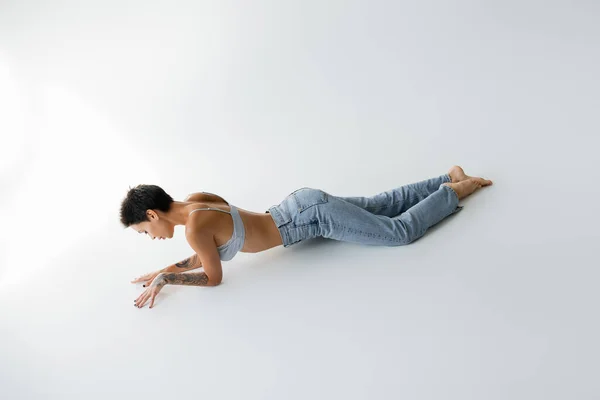 Full length of sexy barefoot woman in blue jeans and bra lying on grey background — Stock Photo