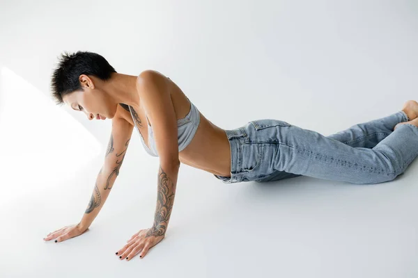 Sensual tattooed woman in satin bra and blue jeans posing on grey background — Photo de stock