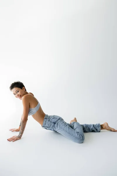 Full length of cheerful barefoot woman in bra and jeans looking at camera while posing on grey background — Fotografia de Stock
