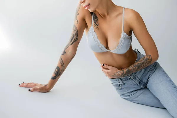 Partial view of woman with sexy tattooed body sitting in jeans and bra on grey background — Photo de stock