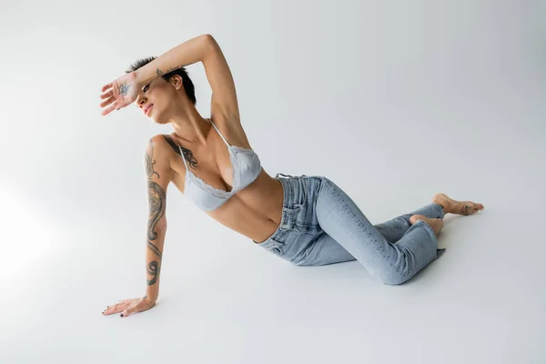 Full length of sexy barefoot woman in silk bralette and blue jeans obscuring face with hand while posing on grey background — Photo de stock