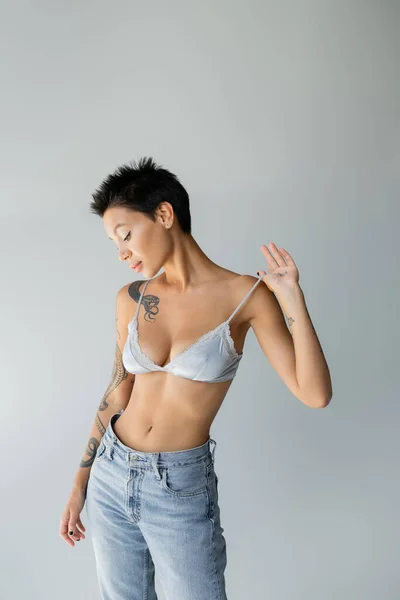 Seductive brunette woman touching strap of blue bralette while posing in blue jeans isolated on grey — Photo de stock