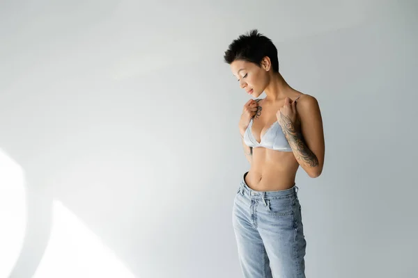 Sexy woman with short brunette hair wearing blue jeans and touching straps of satin bra on grey background — Foto stock