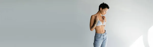 Slender tattooed woman in blue jeans touching straps of silk bralette on grey background, banner — Photo de stock