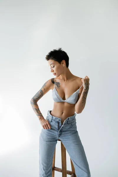 Tattooed woman in unzipped jeans and silk bra posing near high stool on grey background — Photo de stock