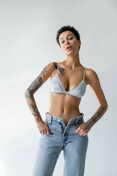 Seductive woman in blue silk bra posing with thumbs in pockets of unzipped jeans while looking at camera on grey background — Photo de stock