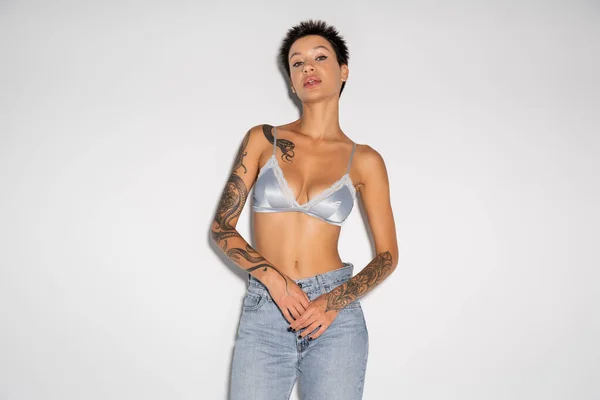 Front view of brunette tattooed woman in silk bra and blue jeans looking at camera on grey background — Stock Photo
