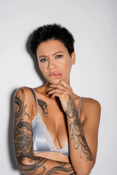 Displeased tattooed woman in blue satin bra biting lip and touching chin while looking at camera on grey background — Stock Photo