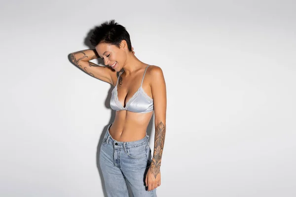 Smiling and sexy woman in silk bralette and jeans standing with hand behind head on grey background — Photo de stock