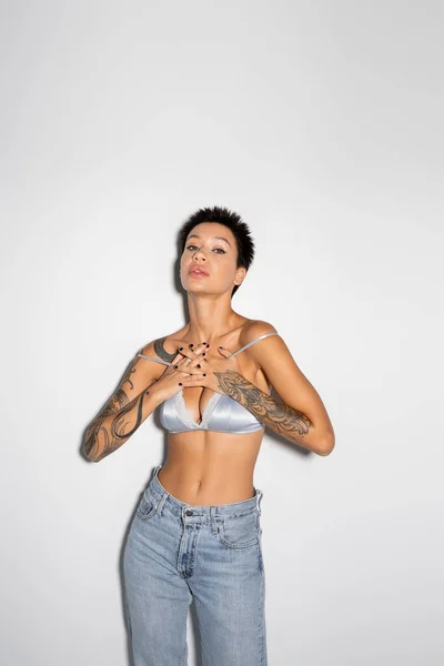 Sexy tattooed woman in silk bra and jeans holding hands on chest while looking at camera on grey background — Foto stock