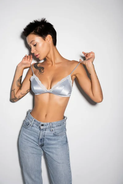 Sexy woman with short brunette hair touching straps of silk bra while standing in jeans on grey background — Photo de stock