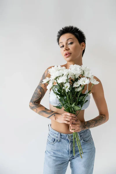Брюнетка татуйована жінка в бюстгальтері і джинсах тримає букет з білих квітів і дивиться на камеру ізольовано на сірому — стокове фото