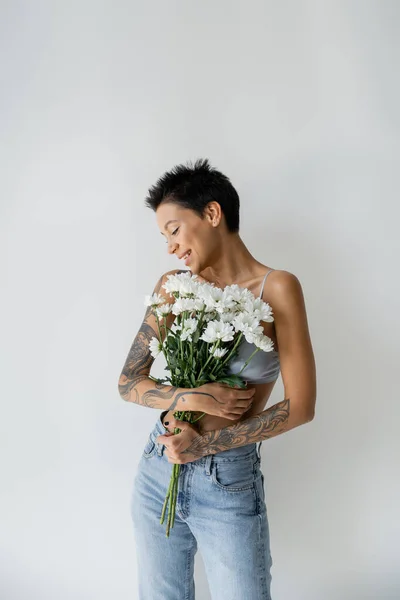 Felice donna tatuata in reggiseno e jeans con bouquet di fiori bianchi isolati su grigio — Foto stock