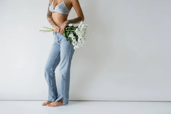 Cropped view of barefoot woman in jeans and bra holding white flowers on grey background — Fotografia de Stock