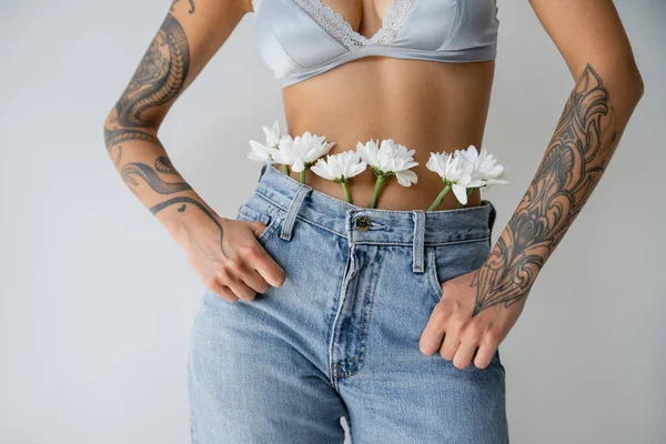 Vista ritagliata di donna tatuata in bralette di seta e jeans con fiori bianchi in piedi con i pollici in tasche isolate su grigio — Foto stock