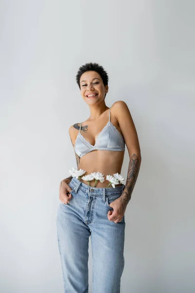 Cheerful tattooed woman in silk bra and white chrysanthemums in jeans smiling at camera isolated on grey — Stockfoto