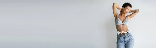 Mulher tatuada feliz vestindo sutiã azul e jeans com flores brancas de mãos dadas atrás do pescoço isolado em cinza, banner — Fotografia de Stock