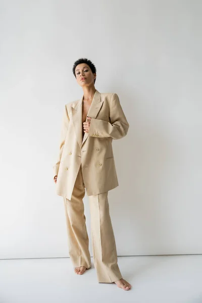Full length of barefoot woman in elegant oversize suit standing and looking at camera on grey background — Stock Photo